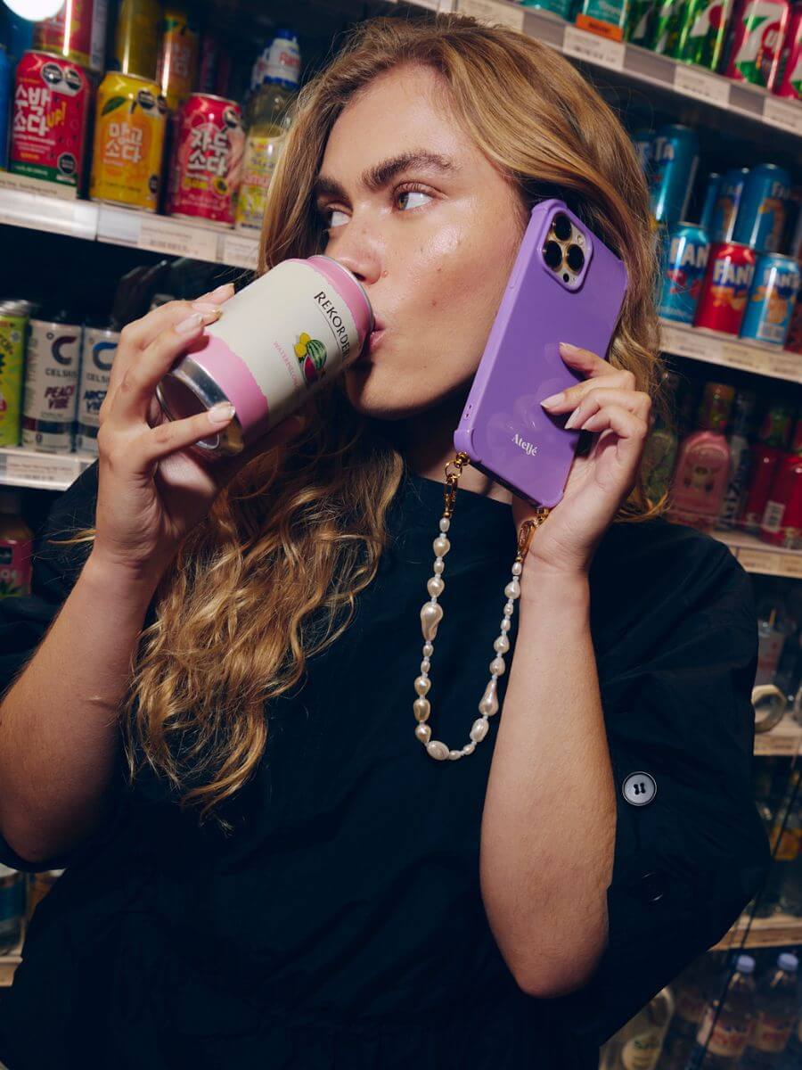 Atelje iphone case violet with pearly Balance short cord, held to model wearing navy jumper's ear in supermarket