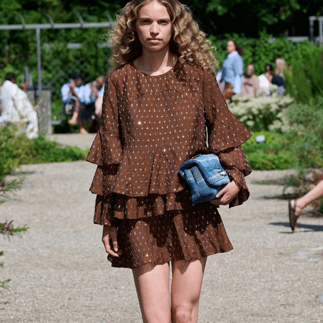 Model on outdoor catwalk in brown Munthe mini dress holding clutch bag