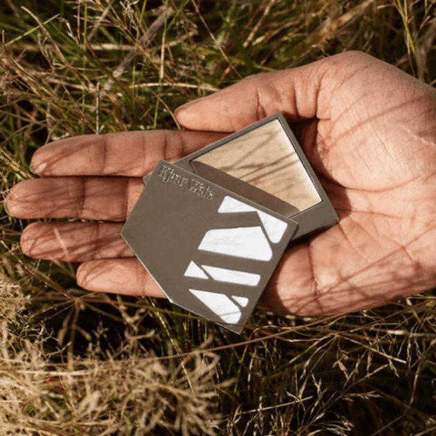 Kjaer Weis Powder in silver case held in palm of hand laying on dry grass in sunshine