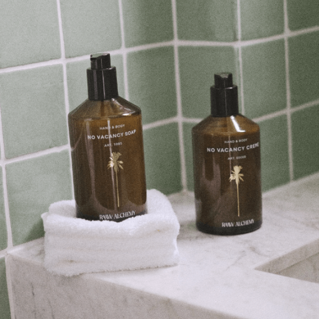 RAAW Alchemy No Vacancy Soap and Creme sitting on the edge of a white marble bath tub with white towel against sage green wall tile